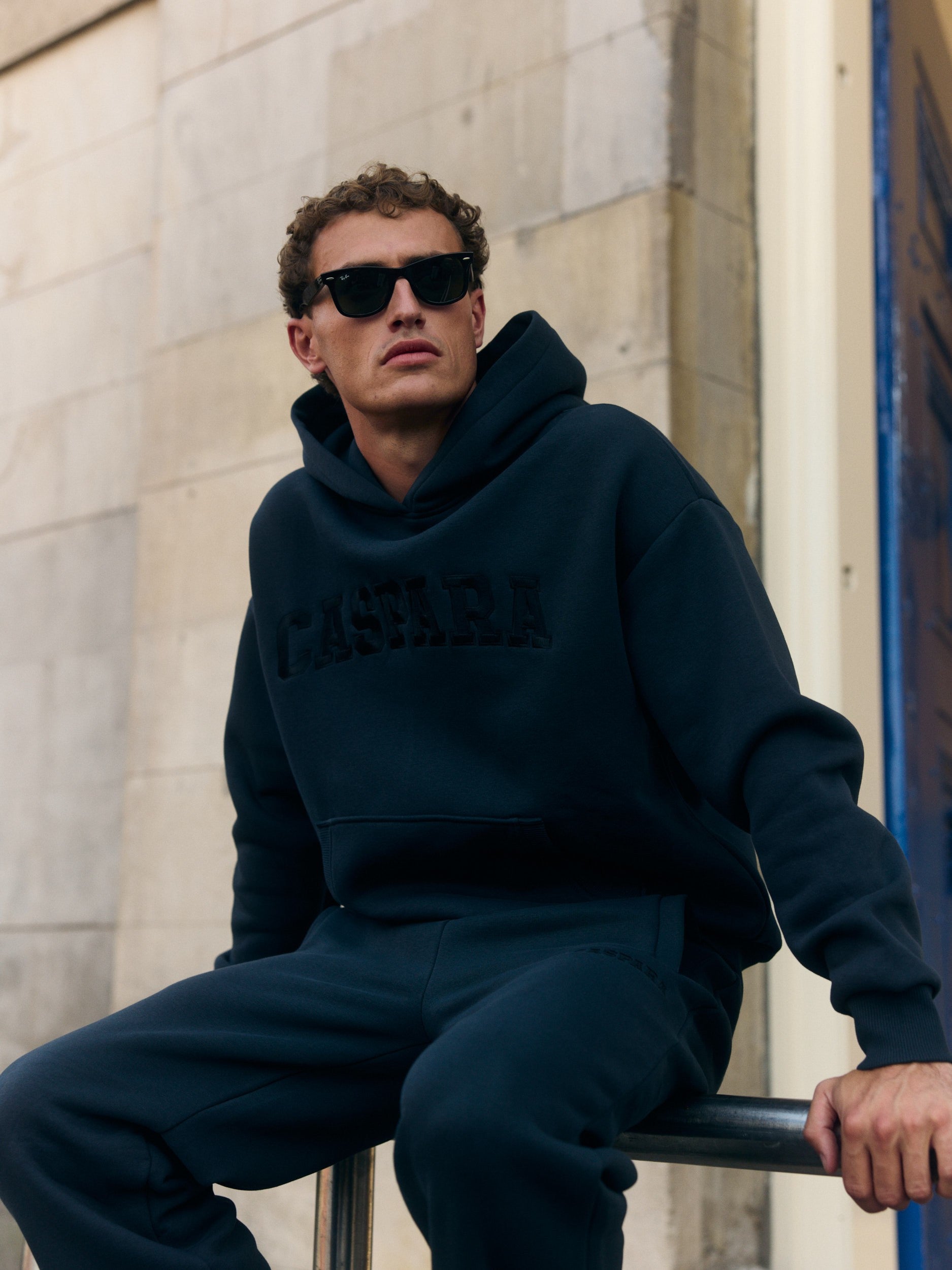Man wearing a black CASPARA hoodie and sunglasses sitting on a railing.